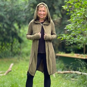 May include: An olive green hooded cardigan with a button closure. The long-sleeved cardigan is worn over a black top and black leggings. The person is standing in a grassy area with trees in the background.