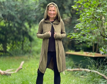 Op de afbeelding: Een olijfgroene cardigan met capuchon en knoopsluiting. De cardigan met lange mouwen wordt gedragen over een zwarte top en zwarte legging. De persoon staat in een grasveld met bomen op de achtergrond.