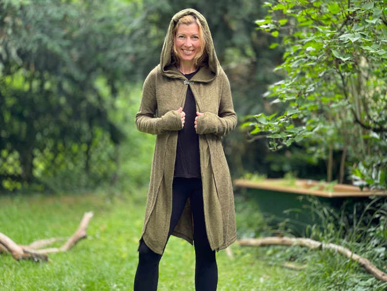 May include: An olive green hooded cardigan with a button closure. The long-sleeved cardigan is worn over a black top and black leggings. The person is standing in a grassy area with trees in the background.