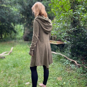 May include: A long-sleeved olive green hooded coat with a flared skirt, paired with black leggings. The coat has a hood and a button closure. The person is standing barefoot on green grass, surrounded by trees.