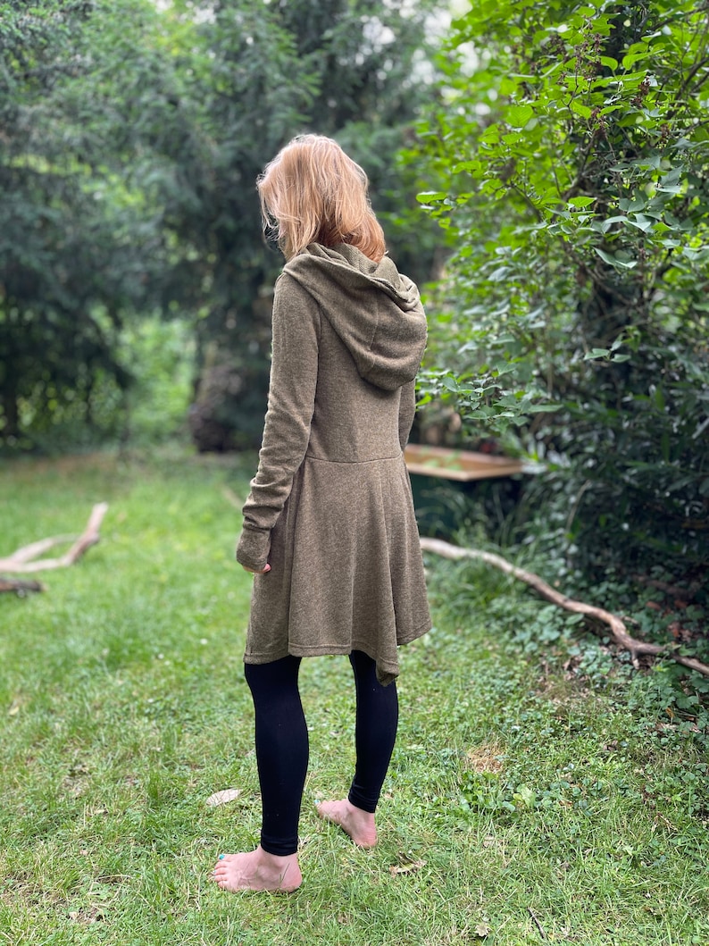 May include: A long-sleeved olive green hooded coat with a flared skirt, paired with black leggings. The coat has a hood and a button closure. The person is standing barefoot on green grass, surrounded by trees.