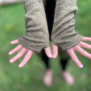 May include: Olive green knit fingerless gloves with ribbed cuffs. The gloves are made of a textured fabric and are shown with open hands against a blurred green background. The sleeves extend to the mid-forearm.