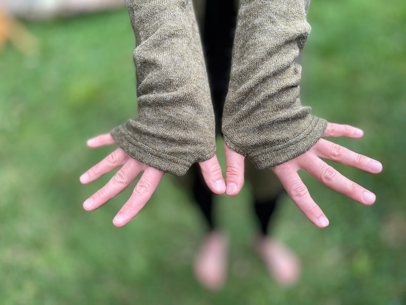 May include: Olive green knit fingerless gloves with ribbed cuffs. The gloves are made of a textured fabric and are shown with open hands against a blurred green background. The sleeves extend to the mid-forearm.