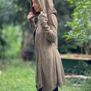 May include: A long olive green hooded cardigan with a flowing, asymmetrical hem. The cardigan has long sleeves and a deep hood. The person in the image is wearing black leggings. The background is a blurred green outdoor scene.