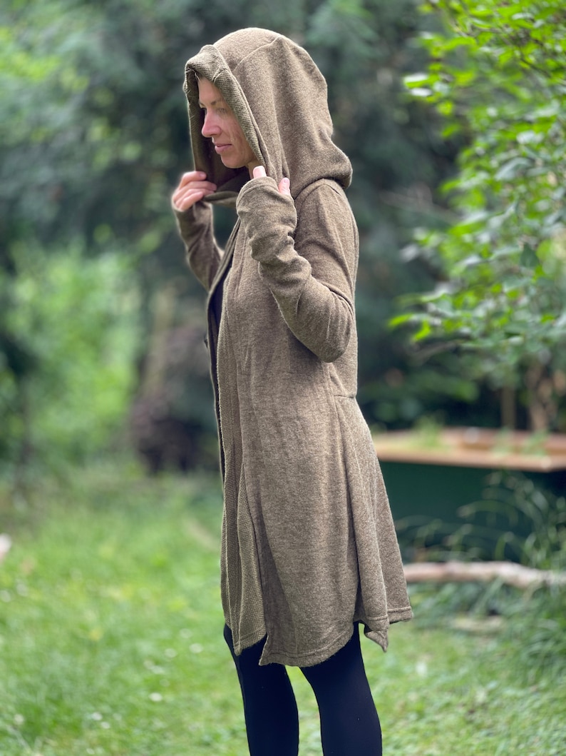 May include: A long olive green hooded cardigan with a flowing, asymmetrical hem. The cardigan has long sleeves and a deep hood. The person in the image is wearing black leggings. The background is a blurred green outdoor scene.
