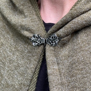 May include: A silver-colored cloak clasp with an ornate, swirling design. The clasp is attached to a hooded, olive-green knitted garment. The clasp is centered on the garment, and the hood is folded over.