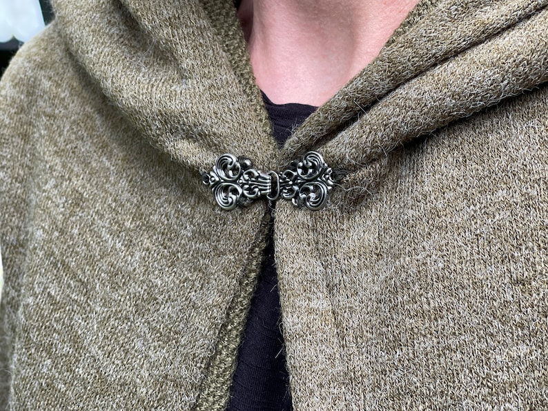 May include: A silver-colored cloak clasp with an ornate, swirling design. The clasp is attached to a hooded, olive-green knitted garment. The clasp is centered on the garment, and the hood is folded over.