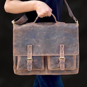 May include: A brown leather briefcase with two front pockets and a long strap. The briefcase has brass buckles and stitching.