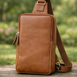 May include: A brown leather sling bag with a rectangular shape and a single adjustable strap. The bag features a zippered main compartment and a smaller zippered pocket. The hardware is gold-colored.