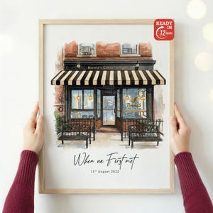 May include: Watercolor print of "Norma's Corner Shoppe" with a black and white striped awning. The print includes the text "When we First met" and "11th August 2022". The print is in a wooden frame and is ready in 12 hours.