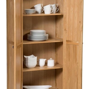 Puede incluir: Un armario de madera con dos puertas, abierto para revelar estantes llenos de platos y tazas blancas. El armario est&aacute; hecho de madera clara y tiene un dise&ntilde;o sencillo.