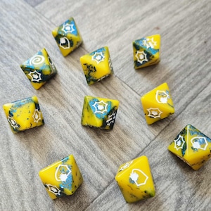 May include: A collection of yellow, eight-sided dice with blue and black marbling. Each die features white geometric designs. The dice are scattered on a grey wooden surface, creating a visually appealing arrangement.