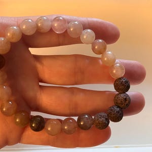 May include: A beaded bracelet featuring a series of smooth, translucent, light-colored stone beads in shades of peach, pink, and tan, transitioning into a section of four dark, porous, textured volcanic rock beads arranged in a circular pattern.