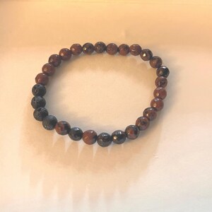 May include: A circular stretch bracelet composed of alternating beads. The majority of the beads are faceted, polished brown stones with a reflective surface, while a small section consists of three matte, porous black volcanic rock beads.