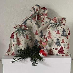 Könnte beinhalten: Drei Weihnachtsgeschenktüten aus Stoff mit Kordelverschlüssen. Die Taschen zeigen ein sich wiederholendes Muster aus roten, grauen und weißen Weihnachtsbäumen und Sternen. Ein Holzvogel-Ornament und eine rot-weiße Kugel sind ebenfalls vorhanden.