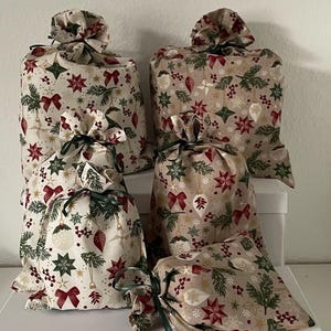 Könnte beinhalten: Mehrere Stoff-Geschenktüten mit festlichem Weihnachtsmuster. Die Beutel sind beige mit roten Schleifen, grünen Zweigen und goldenen Stern- und Ornament-Designs. Jeder Beutel ist mit einem grünen Band gebunden.