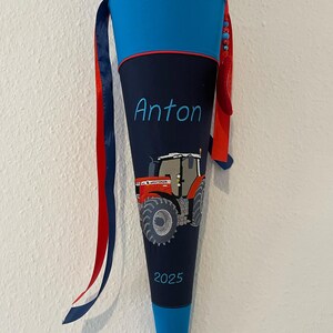 Könnte beinhalten: Eine blaue und marinefarbene Schultüte mit roten und blauen Bändern. Die Tüte zeigt den Namen "Anton" in Schreibschrift, eine rote Traktorgrafik und das Jahr "2025". Der obere und untere Teil sind blau, mit einem marinefarbenen Mittelteil.