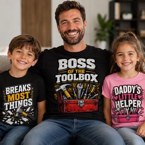 May include: A family of three wearing matching graphic t-shirts. The father's shirt says "BOSS OF THE TOOLBOX" with tools in a toolbox. The son's shirt says "BREAKS MOST THINGS" with a hammer. The daughter's shirt says "DADDY'S LITTLE HELPER".