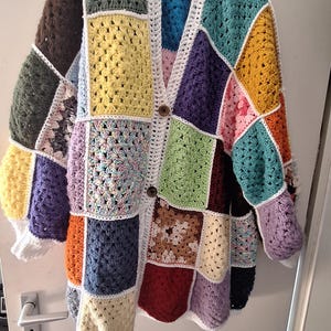 Könnte beinhalten: Ein farbenfroher Patchwork-Häkelcardigan mit V-Ausschnitt und langen Ärmeln. Der Pullover hat verschiedene quadratische Muster in Blau-, Grün-, Gelb-, Lila- und Orangetönen, mit weißer Einfassung und zwei braunen Knöpfen.
