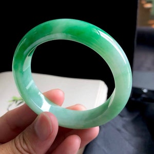 May include: A green jade bangle bracelet with a smooth, polished finish. The bracelet is a light green color with a white undertone.