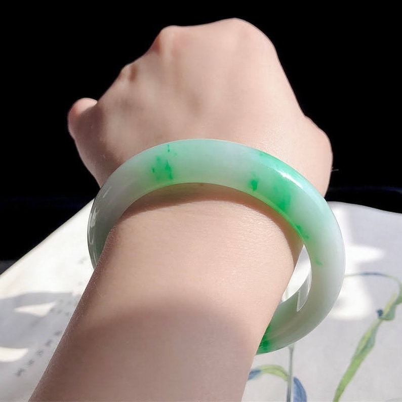 May include: A jade bangle bracelet with a white background and green jade accents. The bangle is smooth and polished.