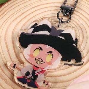 Puede incluir: Un llavero acrílico transparente con un personaje de dibujos animados con un sombrero negro y una sonrisa traviesa. El llavero está sujeto a un cierre plateado y descansa sobre una rodaja de madera, rodeado de tulipanes rosas y naranjas.