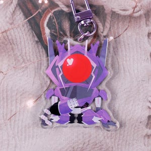 May include: A purple and white anime-style keychain featuring a robot design with a red orb. The keychain has a silver clasp and is hanging against a textured, off-white background. The robot has a geometric design.