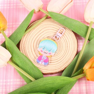 May include: A clear acrylic charm featuring a cartoon character with blue hair, a pink outfit, and a small heart. The charm is on a wooden slice, surrounded by green leaves and tulips in shades of yellow, orange, and cream, set against a pink plaid background.