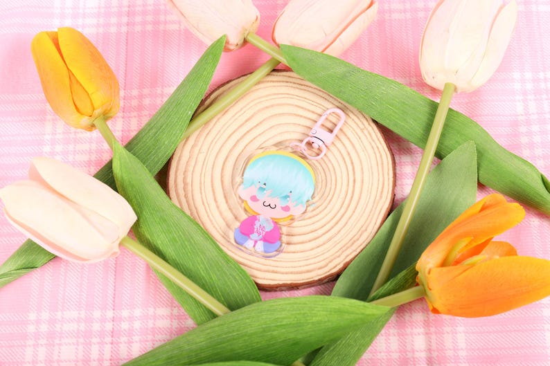 May include: A clear acrylic charm featuring a cartoon character with blue hair, a pink outfit, and a small heart. The charm is on a wooden slice, surrounded by green leaves and tulips in shades of yellow, orange, and cream, set against a pink plaid background.