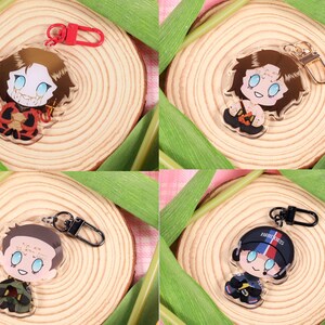 May include: Four acrylic keychains with cartoon character designs. Each keychain features a different character with blue eyes and various outfits. One character wears a red and black outfit, another a black and orange outfit. The keychains are displayed on wooden slices.