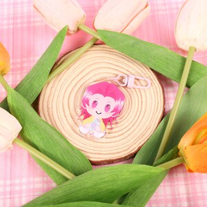 May include: A clear acrylic charm featuring a cartoon character with pink hair and a yellow and white outfit. The charm is attached to a pink keychain. The charm is surrounded by yellow and white tulips and green leaves, set on a pink plaid background.