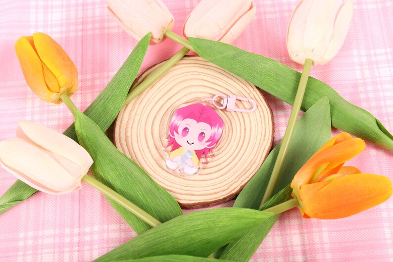 May include: A clear acrylic charm featuring a cartoon character with pink hair and a yellow and white outfit. The charm is attached to a pink keychain. The charm is surrounded by yellow and white tulips and green leaves, set on a pink plaid background.