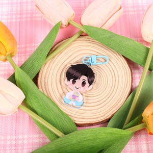 May include: A clear acrylic keychain featuring a cartoon character with black hair and a blue outfit, attached to a light blue clasp. The keychain rests on a wooden slice, surrounded by orange and white tulips with green leaves, set against a pink plaid background.