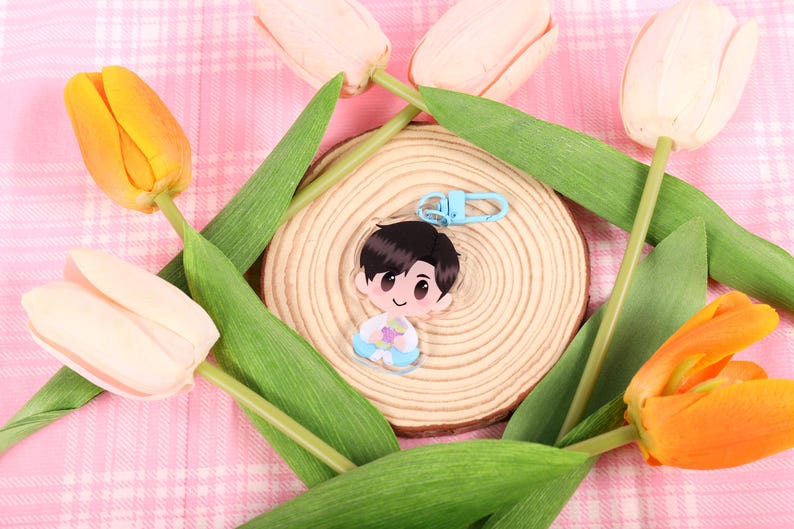 May include: A clear acrylic keychain featuring a cartoon character with black hair and a blue outfit, attached to a light blue clasp. The keychain rests on a wooden slice, surrounded by orange and white tulips with green leaves, set against a pink plaid background.