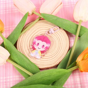 May include: A pink and yellow cartoon character keychain with a pink clasp rests on a wooden slice, surrounded by green leaves and colorful tulips. The background is a pink plaid pattern.