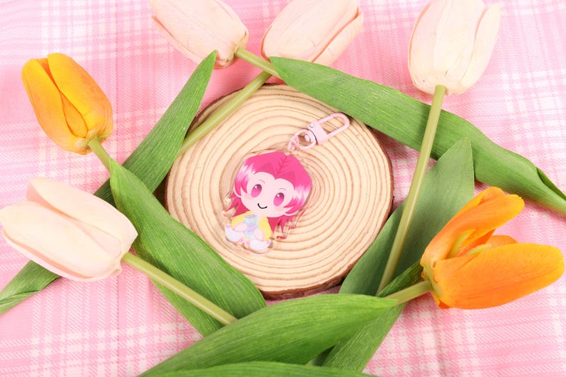 May include: A pink and yellow cartoon character keychain with a pink clasp rests on a wooden slice, surrounded by green leaves and colorful tulips. The background is a pink plaid pattern.