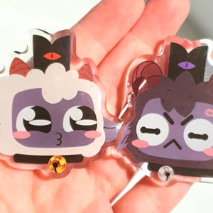 May include: Two acrylic charms featuring cartoon characters with purple and white fur. The characters have large eyes, small mouths, and are wearing black collars with bells. One character has a red crown with a single eye on top, while the other has a purple crown with a single eye on top.