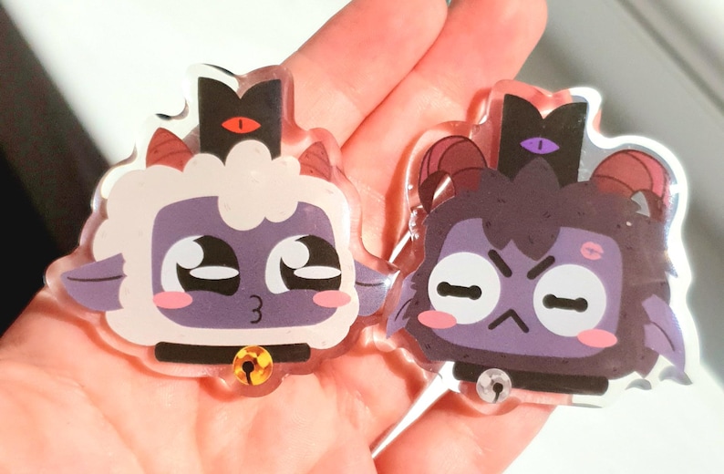 May include: Two acrylic charms featuring cartoon characters with purple and white fur. The characters have large eyes, small mouths, and are wearing black collars with bells. One character has a red crown with a single eye on top, while the other has a purple crown with a single eye on top.