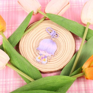 May include: A clear acrylic charm featuring a cartoon character with purple hair and a yellow and purple outfit. The charm is attached to a purple clasp. The charm is on a wooden slice, surrounded by tulips in shades of yellow, orange, and pink.