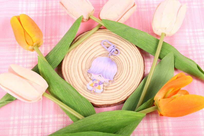 May include: A clear acrylic charm featuring a cartoon character with purple hair and a yellow and purple outfit. The charm is attached to a purple clasp. The charm is on a wooden slice, surrounded by tulips in shades of yellow, orange, and pink.