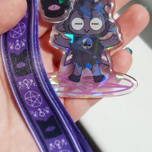 Op de afbeelding: Een paarse lanyard met een herhalend patroon van pentagrammen en symbolen. Aan de lanyard is een holografische hanger bevestigd met een cartoonfiguur met een zwart-wit ontwerp.