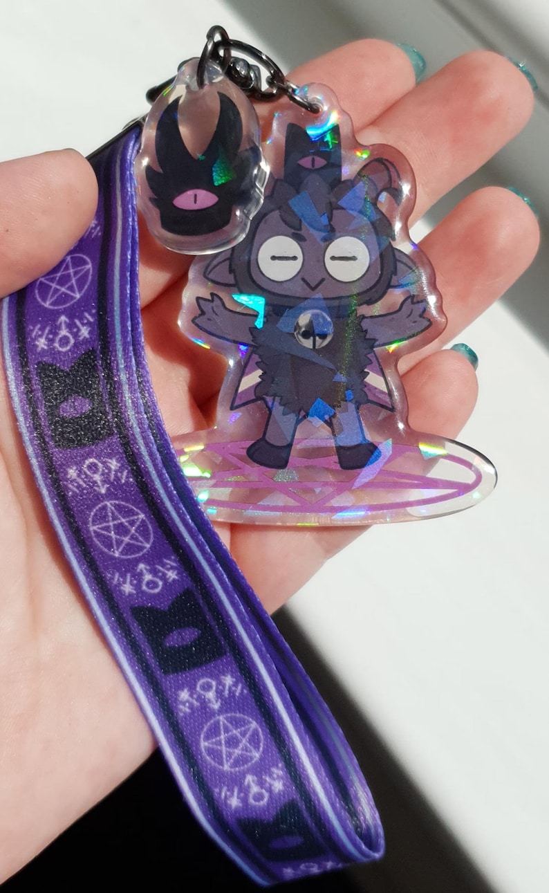 Op de afbeelding: Een paarse lanyard met een herhalend patroon van pentagrammen en symbolen. Aan de lanyard is een holografische hanger bevestigd met een cartoonfiguur met een zwart-wit ontwerp.