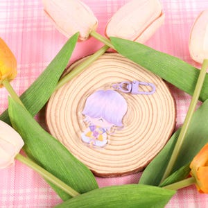 May include: A clear acrylic keychain featuring a cartoon character with lavender hair and a purple clasp. The keychain rests on a wooden slice, surrounded by green leaves and tulips in shades of yellow, orange, and cream, set against a pink plaid background.