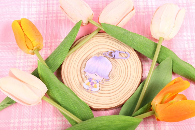 May include: A clear acrylic keychain featuring a cartoon character with lavender hair and a purple clasp. The keychain rests on a wooden slice, surrounded by green leaves and tulips in shades of yellow, orange, and cream, set against a pink plaid background.