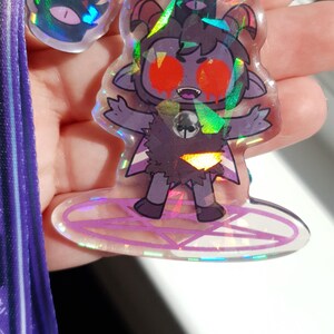 Op de afbeelding: Een holografische sleutelhanger met een cartoonfiguur met rode ogen, hoorns en een pentagram. Het personage draagt een zwart-witte outfit en staat op een pentagram. De sleutelhanger is bevestigd aan een paarse lanyard.