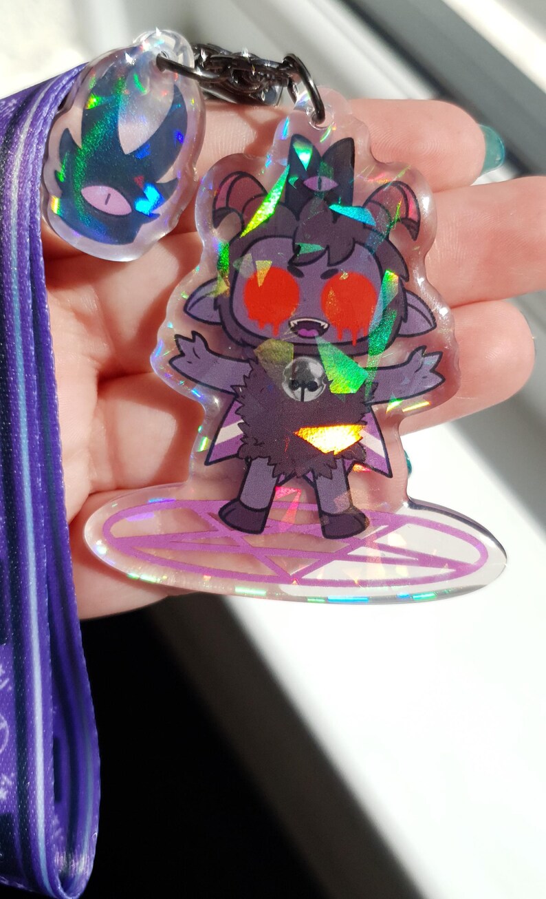 Op de afbeelding: Een holografische sleutelhanger met een cartoonfiguur met rode ogen, hoorns en een pentagram. Het personage draagt een zwart-witte outfit en staat op een pentagram. De sleutelhanger is bevestigd aan een paarse lanyard.