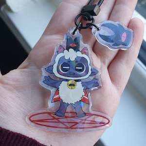 Peut inclure: Un porte-cl&eacute;s en acrylique transparent repr&eacute;sentant un personnage de mouton de dessin anim&eacute; avec une couronne, une cape et une cloche. Le porte-cl&eacute;s a un fermoir argent&eacute; et un plus petit charme avec un personnage sombre. Le mouton est sur un pentagramme rouge.