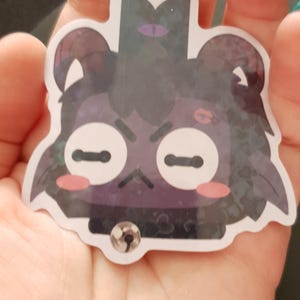 Pu&ograve; includere: Un adesivo di cartone animato nero e viola di un orso con un occhio viola, un rossore rosa e un collare nero con un campanello d'argento. L'adesivo ha una finitura scintillante e glitterata.