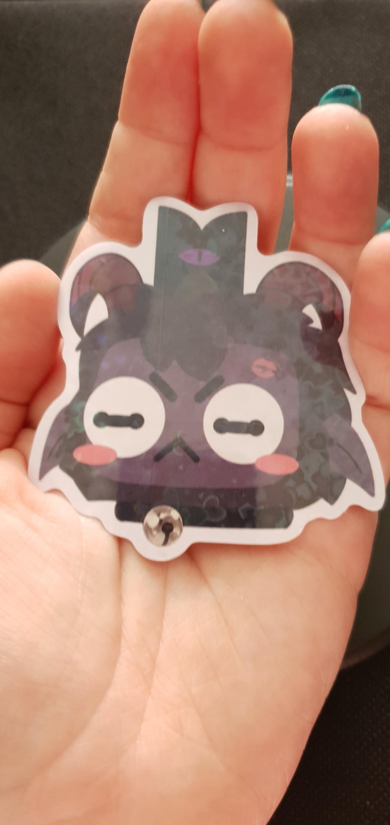 Pu&ograve; includere: Un adesivo di cartone animato nero e viola di un orso con un occhio viola, un rossore rosa e un collare nero con un campanello d'argento. L'adesivo ha una finitura scintillante e glitterata.