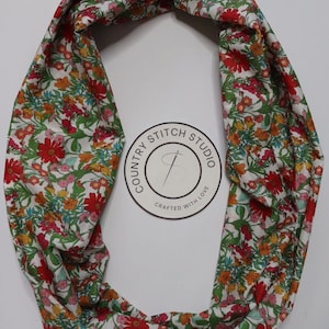 Könnte beinhalten: Ein weißer Stoff-Infinity-Schal mit einem Blumendruck in Rot-, Orange-, Gelb-, Grün- und Blautönen. Der Schal hat ein rundes Logo mit dem Text "Country Stitch Studio Crafted with Love".